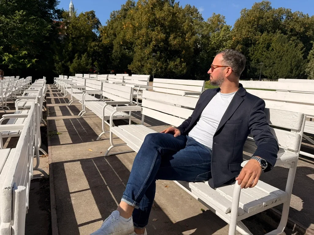 Robert Dobucki w marynarce siedzi na białej ławce w plenerze.